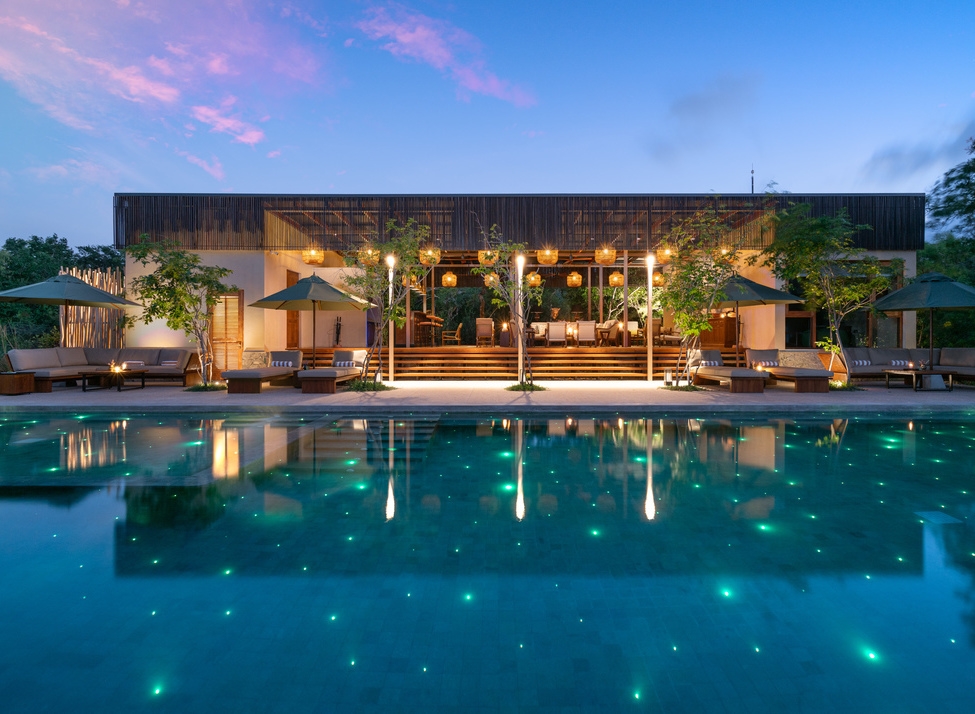 A luxurious infinity pool with glowing green lights at dusk, reflecting a modern pavilion with warm lanterns.
