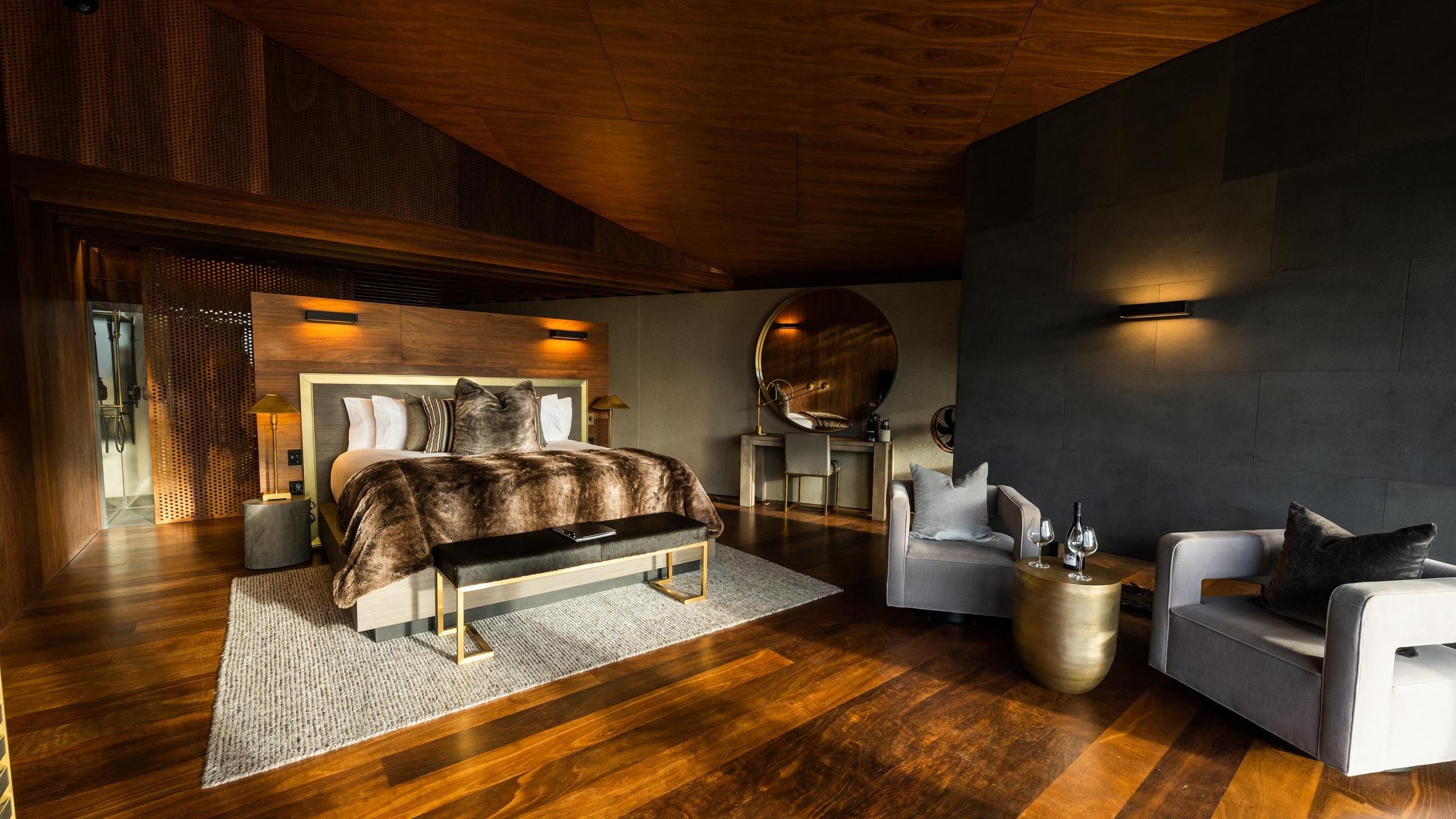 Modern luxury bedroom at Lindis Lodge with wood walls, a large bed with a fur throw, and two grey armchairs.