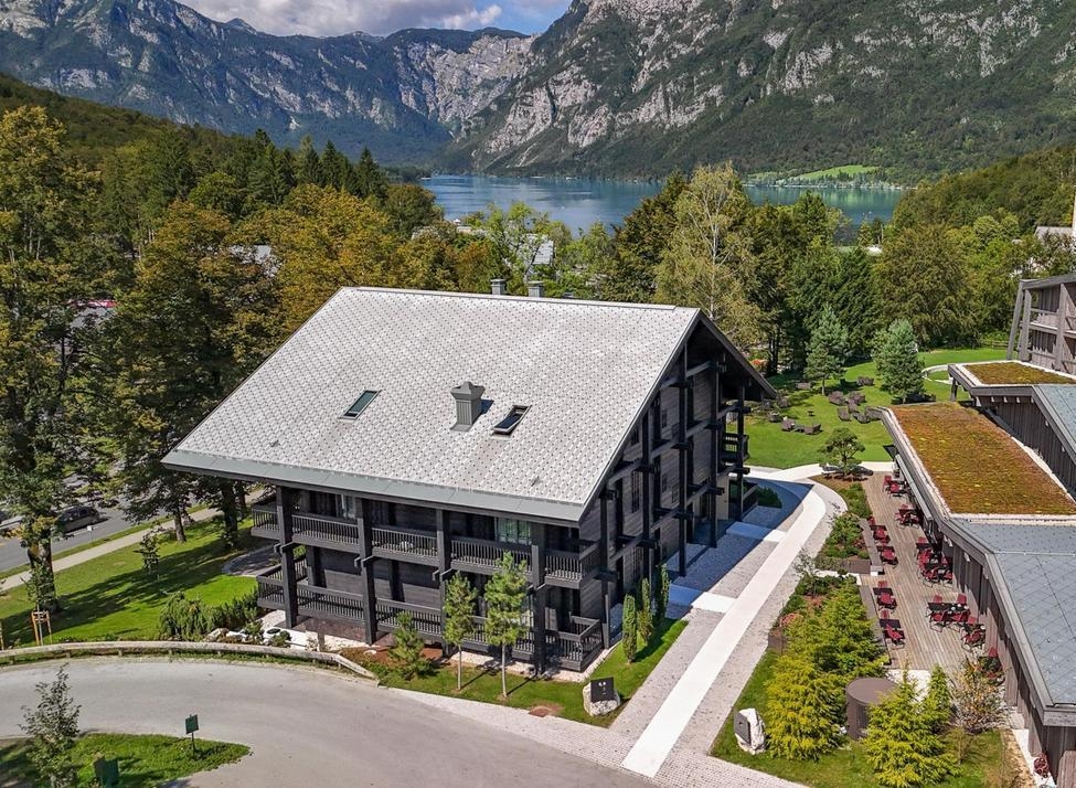 Exterior of Vila Muhr, a multi-story wooden lodge overlooking a blue lake and steep mountain peaks in the distance.