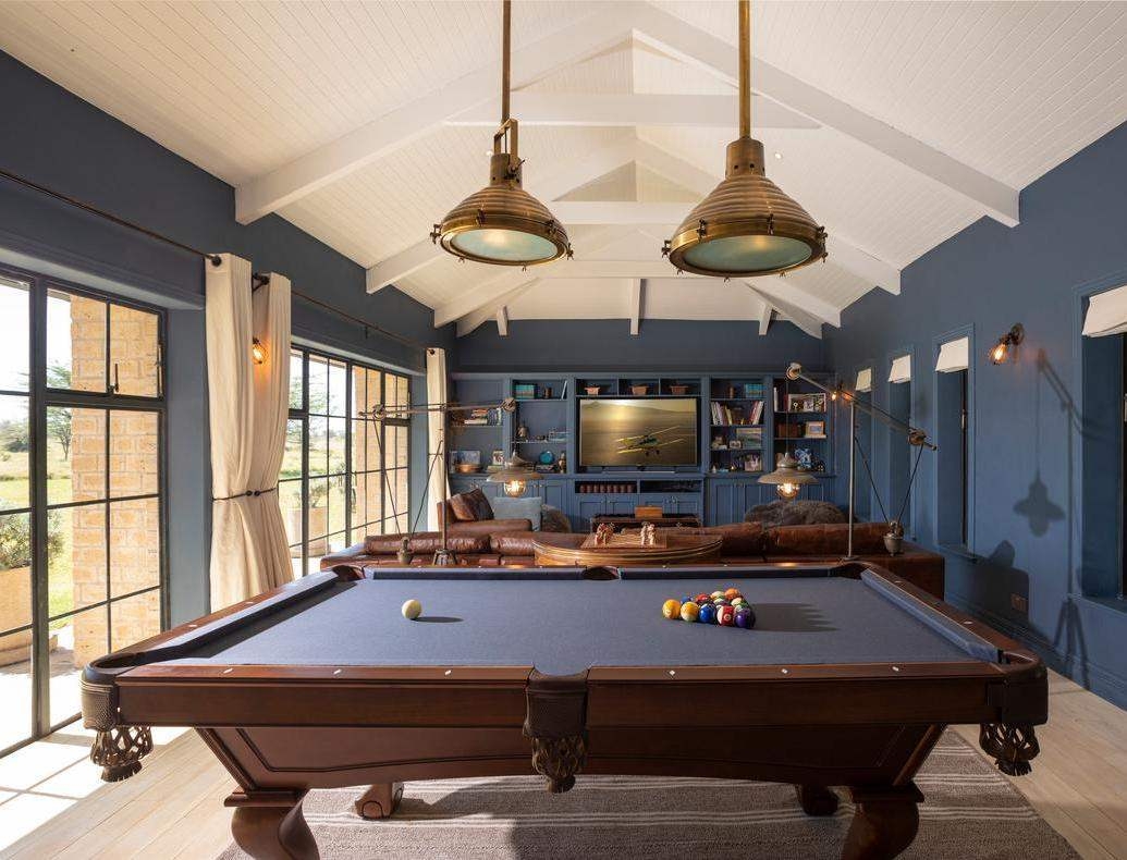 A billiards table in a blue-walled room with brass pendant lights, leather seating and a TV in the background.