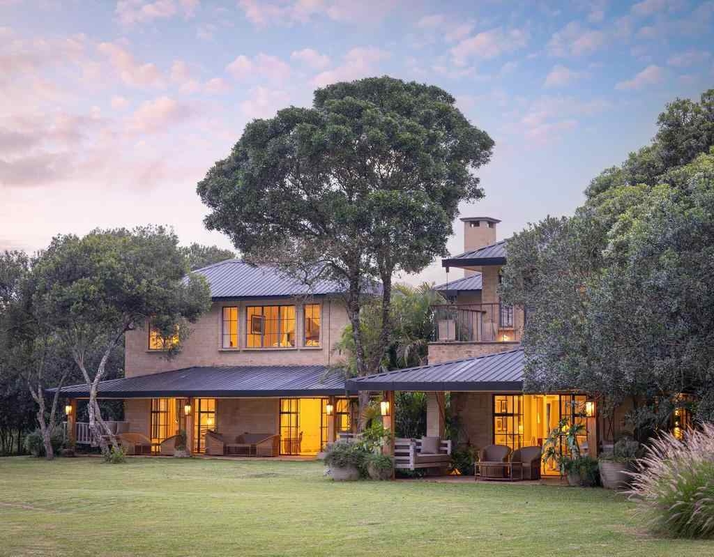 A large two-story house with illuminated windows and a covered patio at dusk surrounded by green trees.