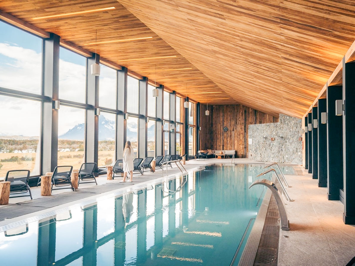 A long thin swimming pool under timber clad ceiling. A woman in a bath robe stands by the pool looking out of the window.