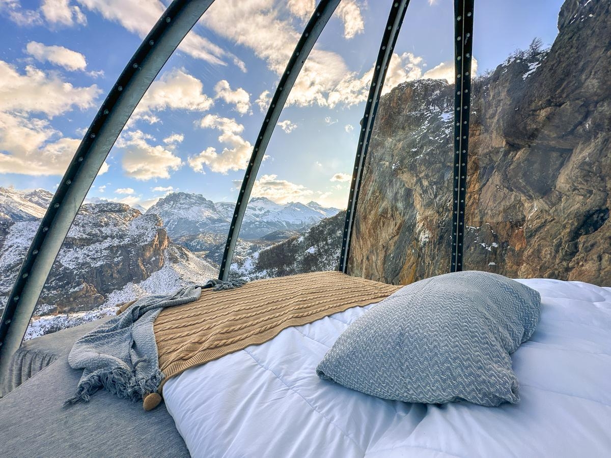 A bed with a grey pillow and tan blanket inside a glass capsule facing a snowy mountain vista in El Chalten.
