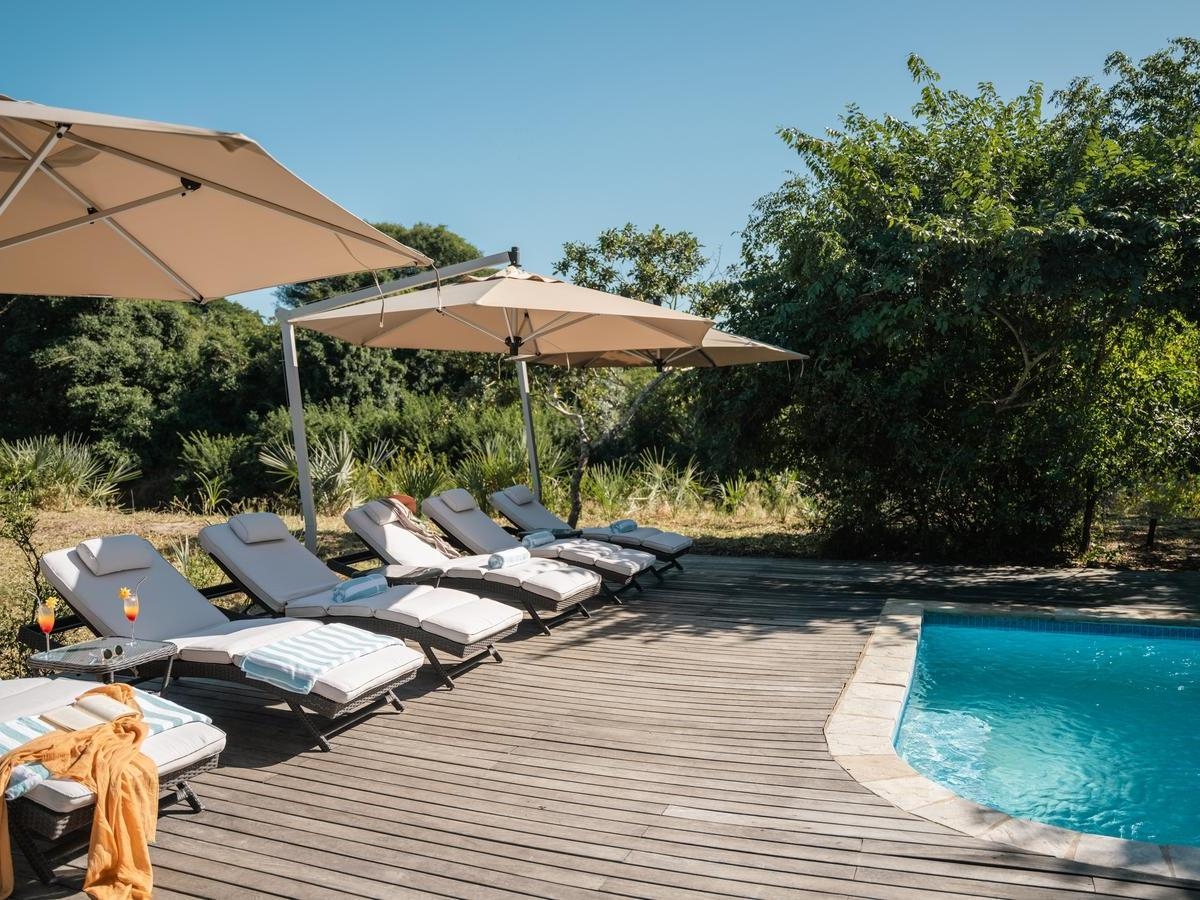 Sun loungers with towels and drinks sit on a wooden deck next to a bright blue swimming pool in a forest.