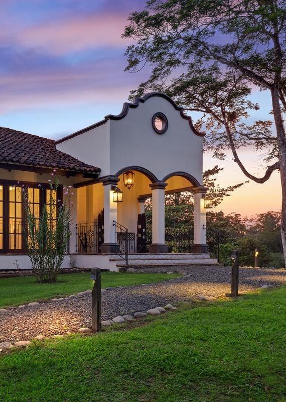Arched entryway of a white house at sunset with glowing lanterns and a gravel path leading to the front.