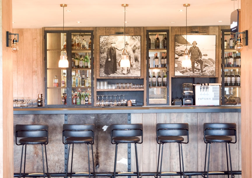 A bar with high chairs in the foreground and shelves with bottles and glasses in the background.