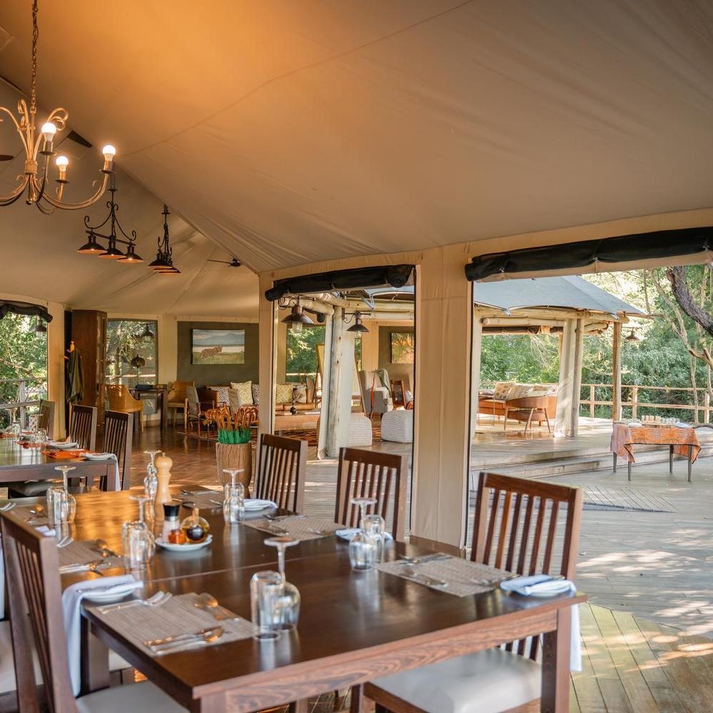 A luxury safari dining room with set wooden tables under a large canvas tent overlooking an outdoor deck.