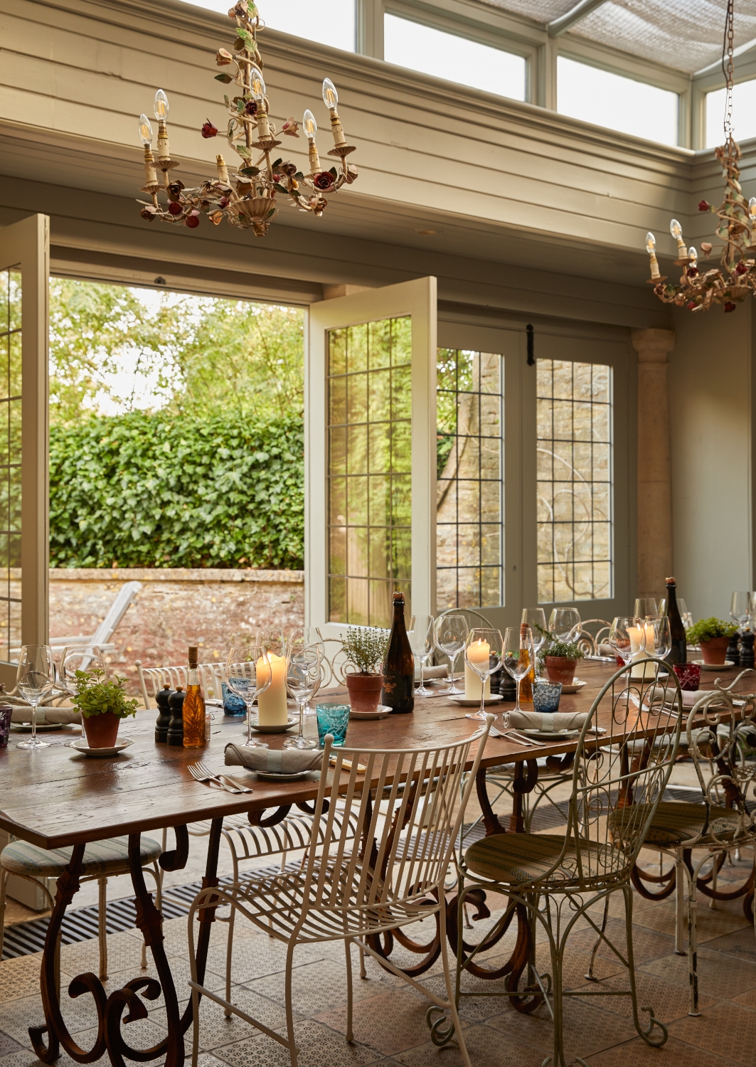 Interior of a long wooden dining table and mismatched chairs in front of double doors leading out to a terrace with greenery.