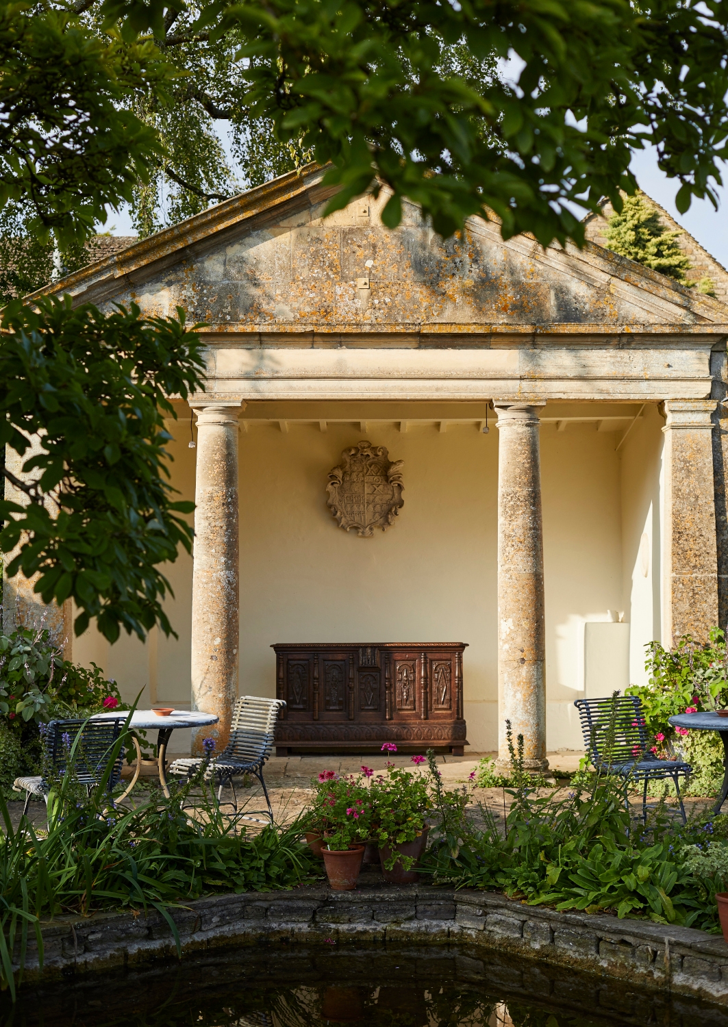 Exterior of a stone building with two stone columsn bathed in sunlight, with a garden table and chairs, and leafy flowers and trees surrounding it.