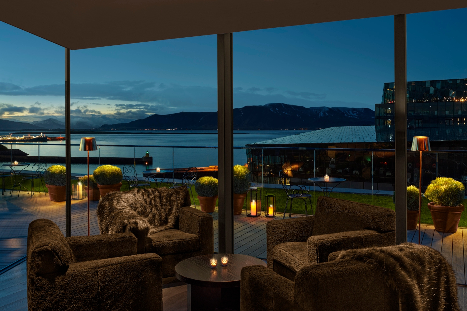 Interior of a bar table with dark grey sofa chairs in a corner with floor-to-ceiling windows looking out over a harbour a dusk.