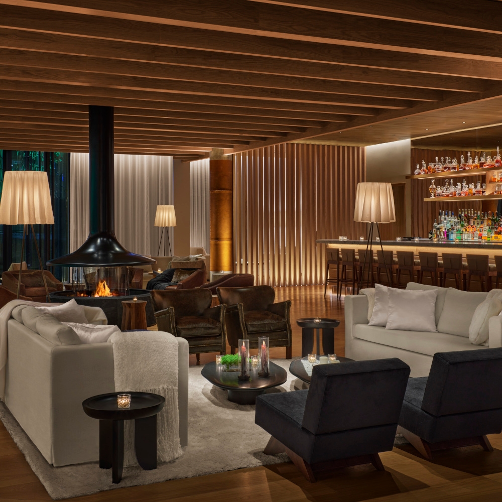 Interior of a bar with white sofa and black chairs, a light wood ceiling and warm lamps, a bar in the background and in the back an open wood stove.