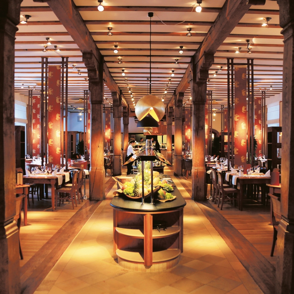 Interior of a hotel restaurant with warm lighting and tall wooden columns and beams, and a buffet counter down the middle of the room with tables on either side.