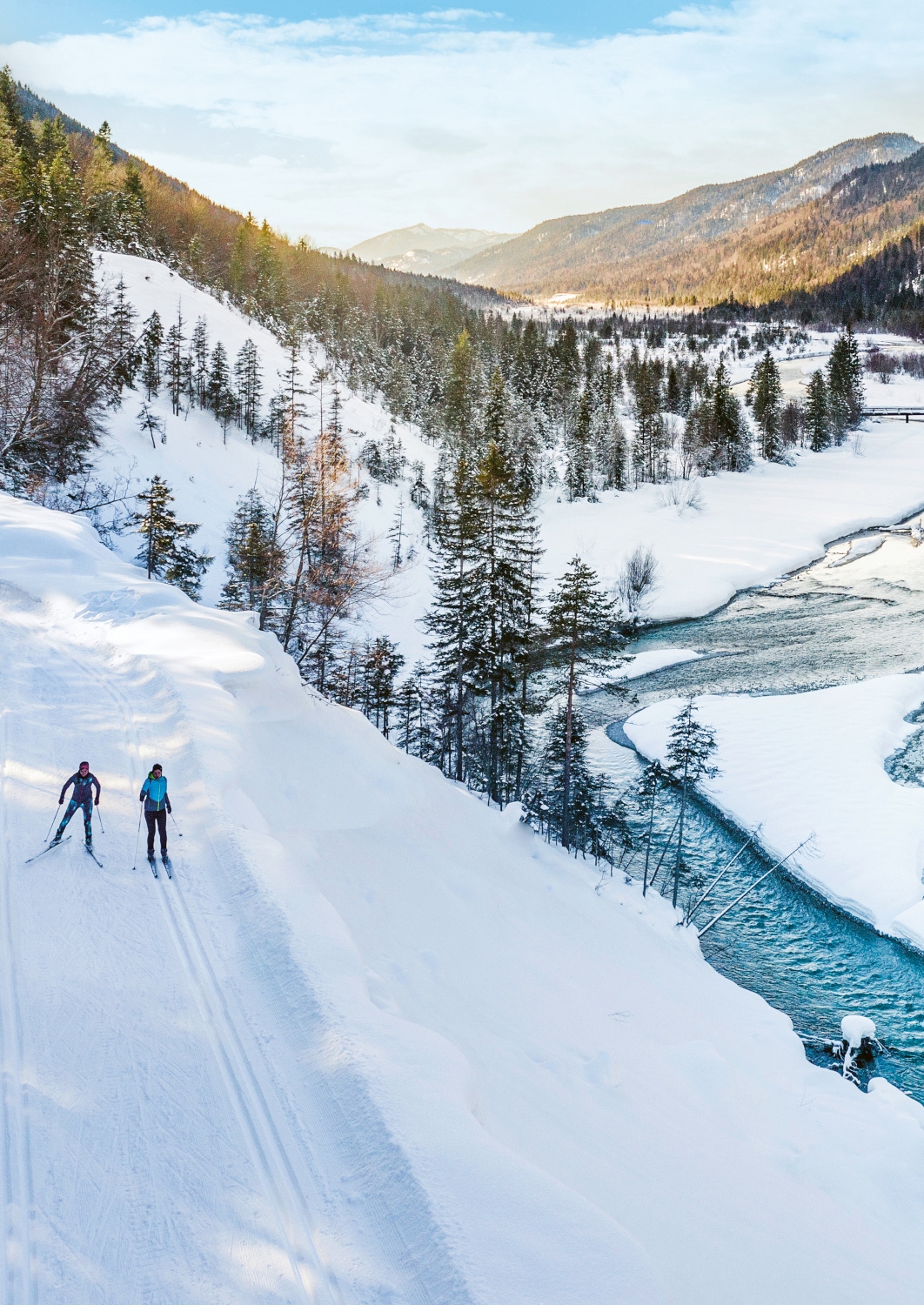 Exterior overhead view of two skiers gliding along a snowy mountainside with a blue river flowing through snow and forest just below them.