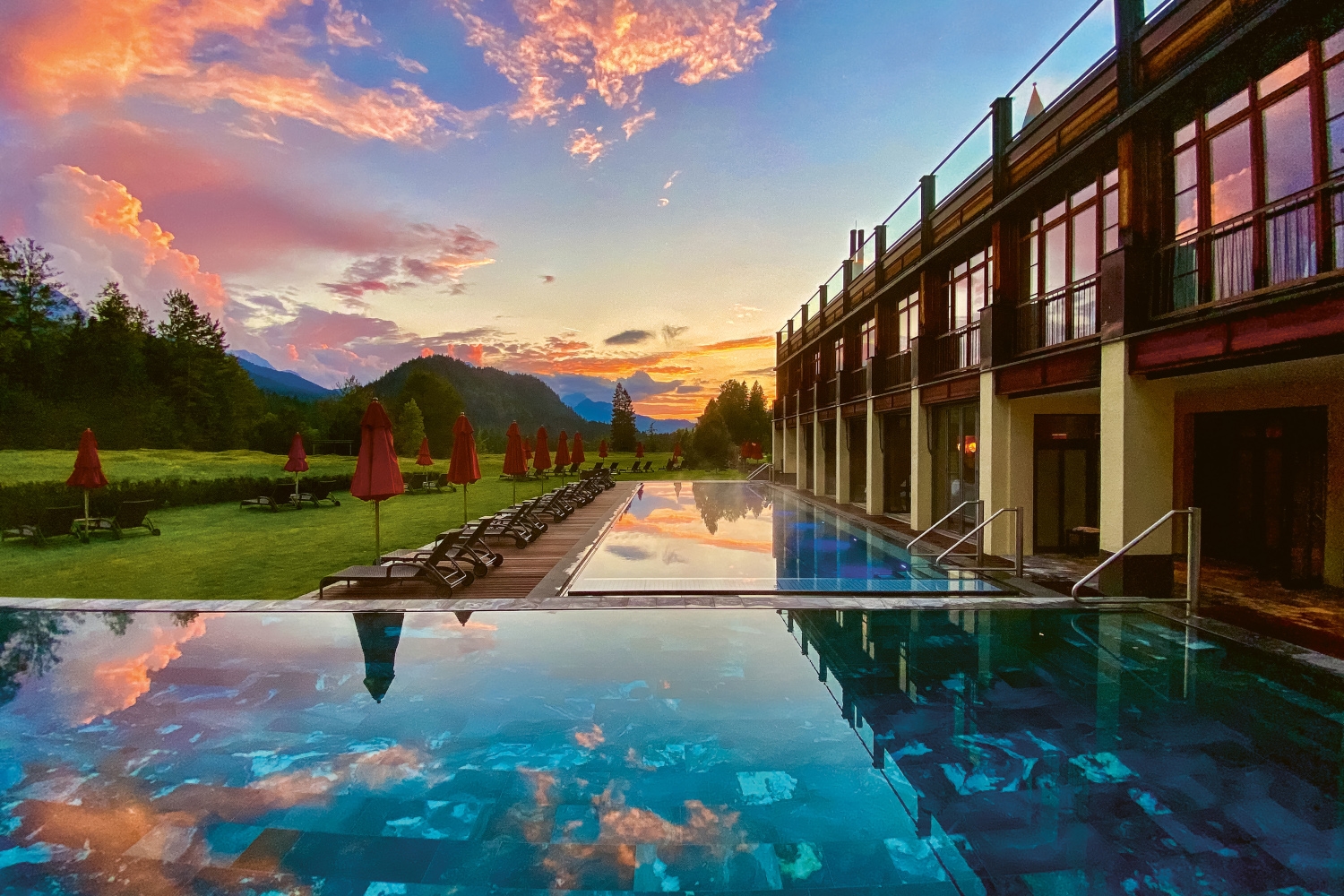 Exterior of a hotel with bright blue swimming pool and mountains in the distance, and a pink and purple sunset sky.