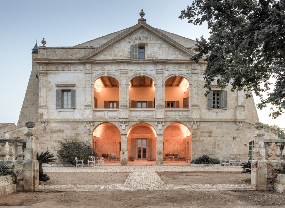 Facade of a historic stone building with atriums lit up inside, and a stone plaza in the foreground with trees on the right side.