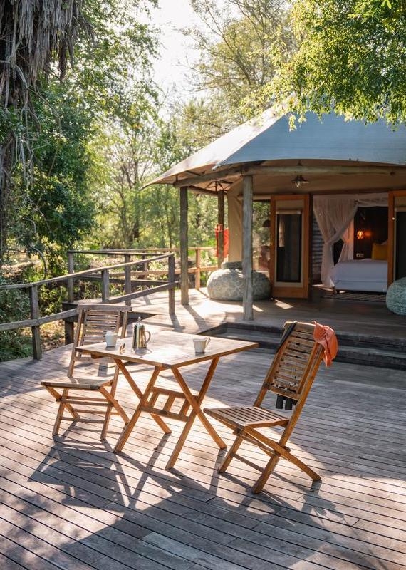 A wooden table and two chairs sit on the deck of a luxury safari tent in the morning sunlight.