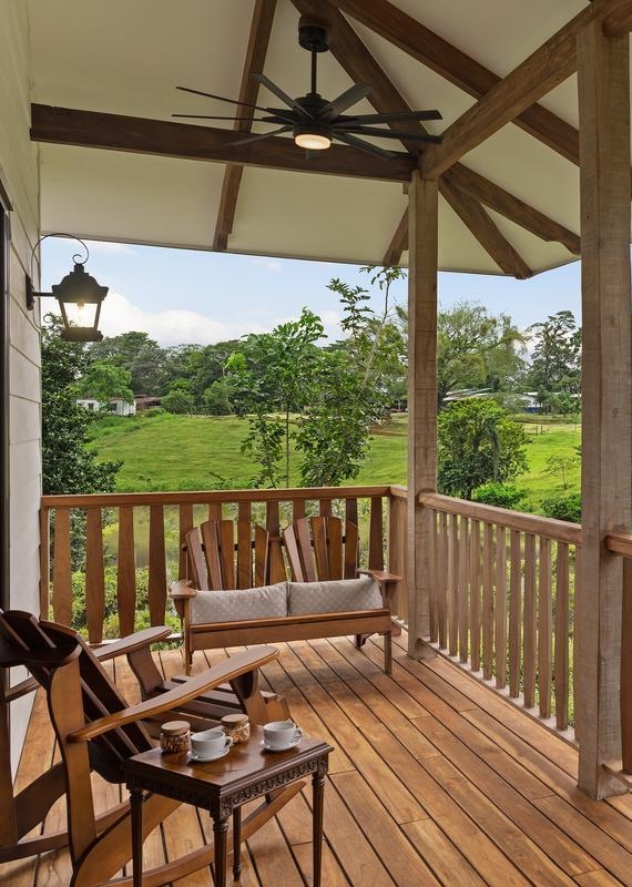 Wooden balcony with rocking chairs and coffee cups under a ceiling fan overlooking green hills and trees.