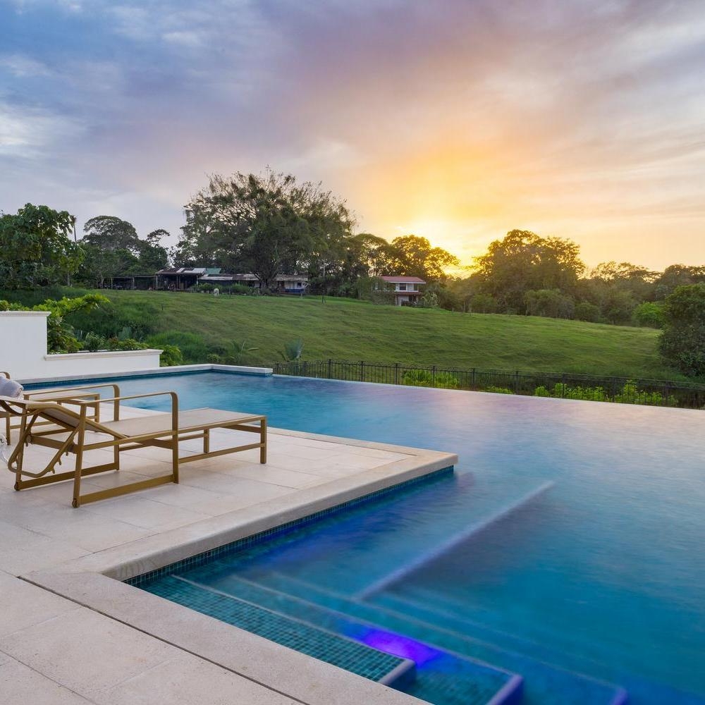 Infinity pool with lounge chairs overlooking a grassy hill and forest under a vibrant orange and purple sunset sky.