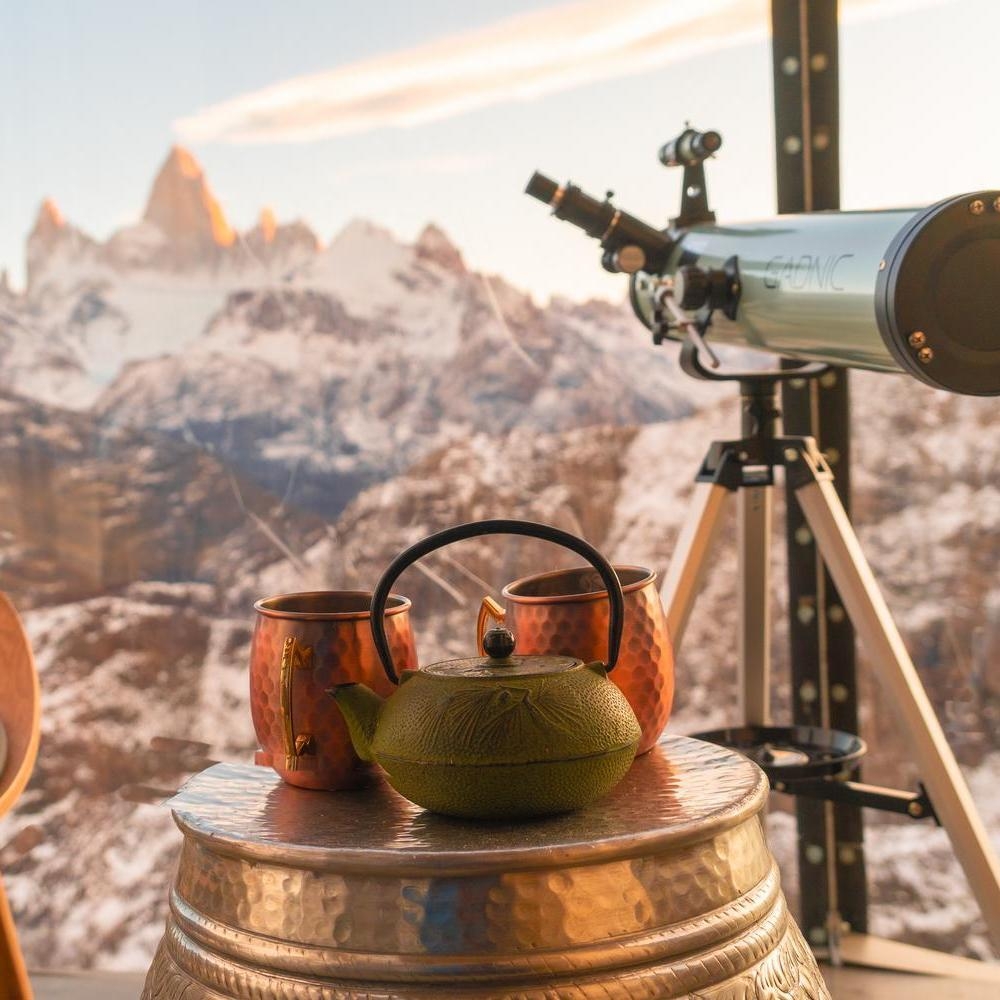 A telescope and green teapot inside an Ovo Patagonia capsule with a view of jagged, snow-capped mountains.
