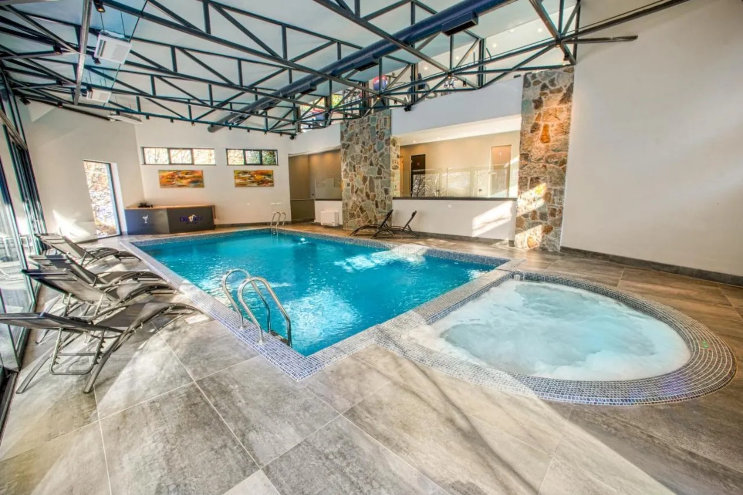 Interior shot of an indoor swimming pool with a Jacuzzi in the foreground, a line of lounge chairs to the left, and above the ceiling is lined with steetl beams.
