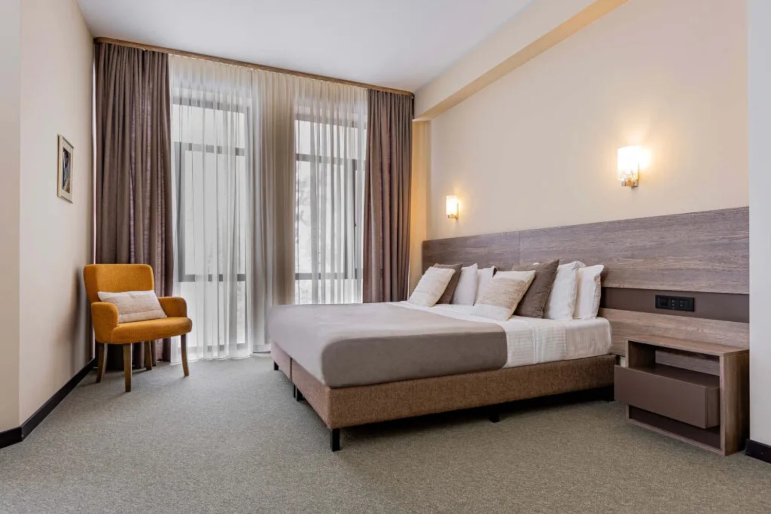 Interior of a Dilijazz Hotel room with a double bed with khaki and white bedding, grey carpet, and a large floor-to-ceiling window with khaki curtains.