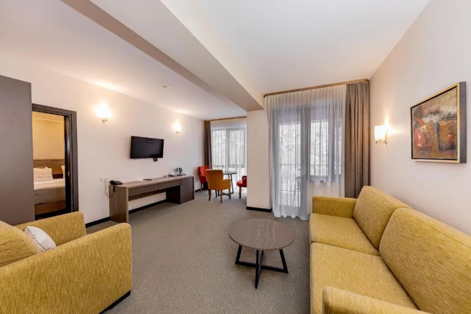 Interior of a Dilijazz Hotel room with a yellow sofa and armchair in in the foreground, a tv on the wall and red chairs in the background, grey carpet, and two large floor-to-ceiling windows with khaki curtains.
