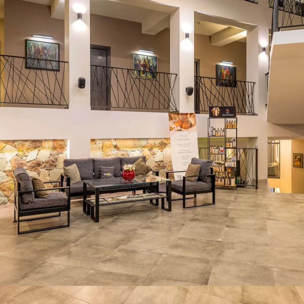 Interior view of the Dilijazz Hotel lobby with beige tile floors and a set of dark grey sofa and chairs in the centre. Above and behind is a line of room doors on a mezzanine level.