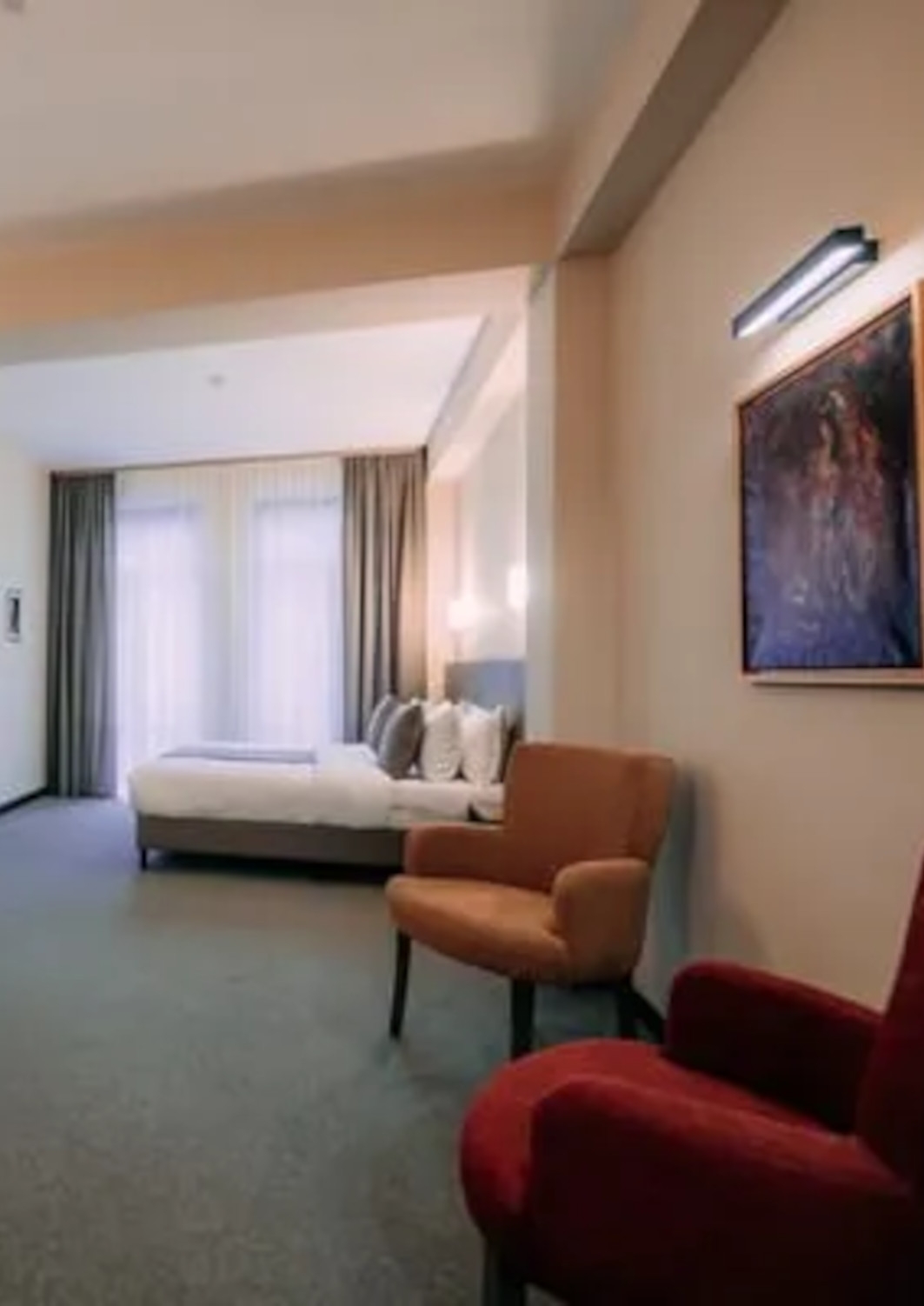 Interior of a Dilijazz Hotel room with a double bed with white bedding, grey carpet, and a large floor-to-ceiling window with khaki curtains. There are two red arm chairs in the foreground.