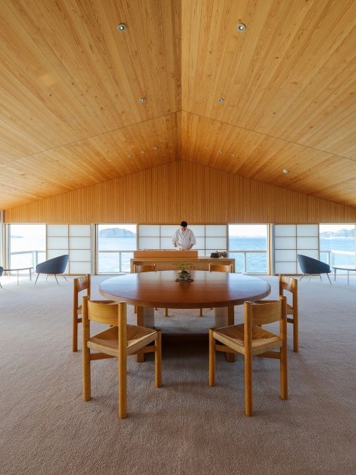 A round wooden table and four chairs under a slanted wooden roof with a chef in the background