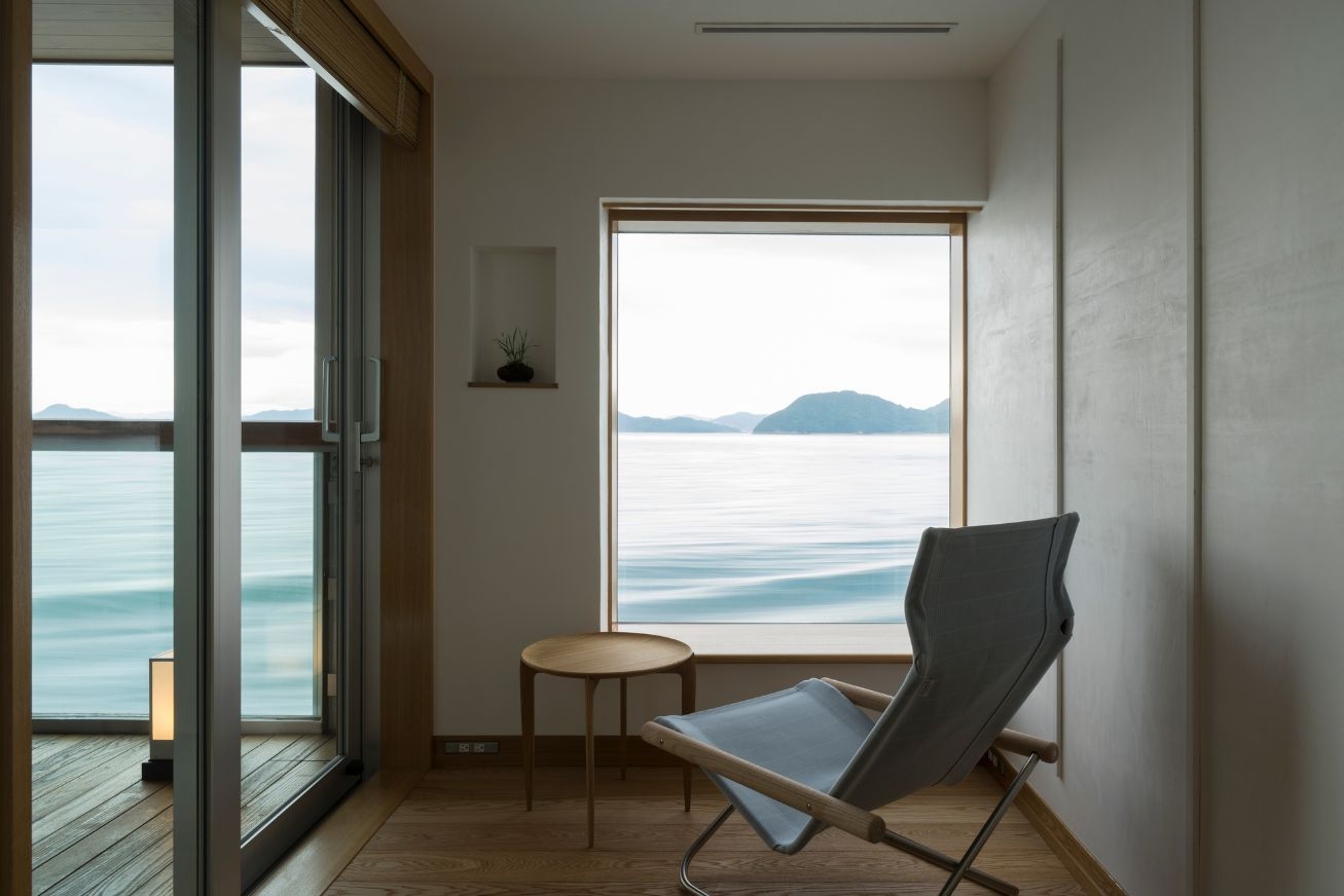 A chair and small wooden table in front of a large window with a view of the sea and mountains