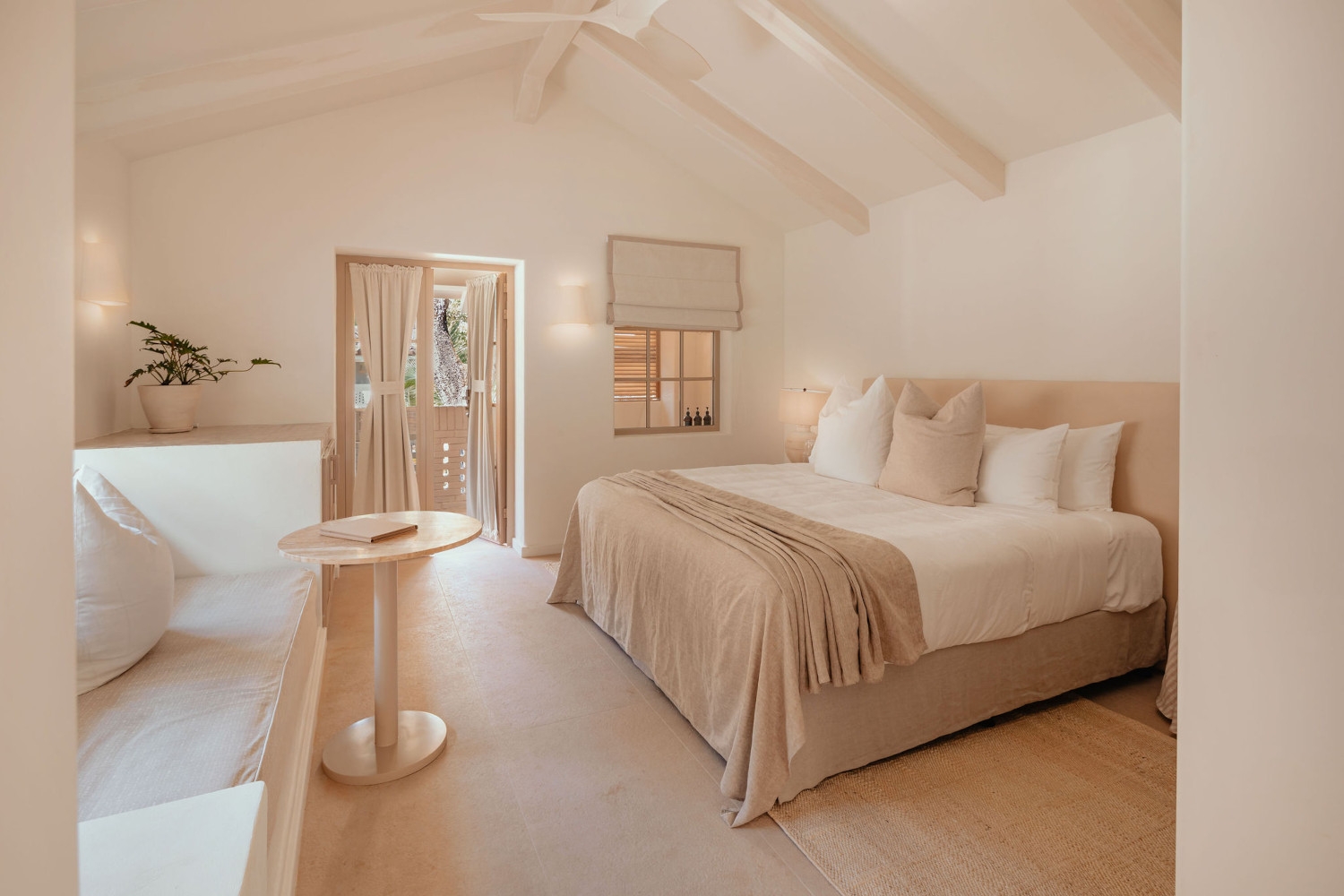 Bedroom interior decorated in beige and white, with a large bed, table, and pitched ceiling and doorway out onto a terrace.