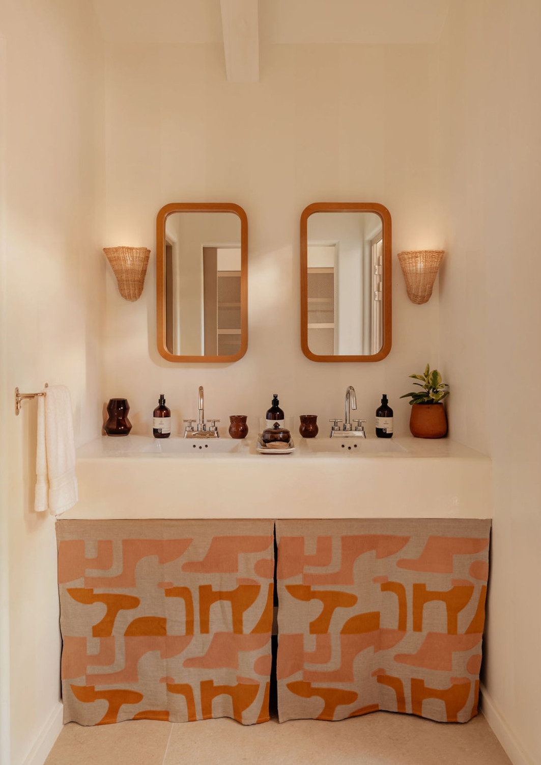 Interior of a bathroom sink with two long mirrors, a white basin and two lamps on either side of them, and assorted toiletries and bottles along the basin.