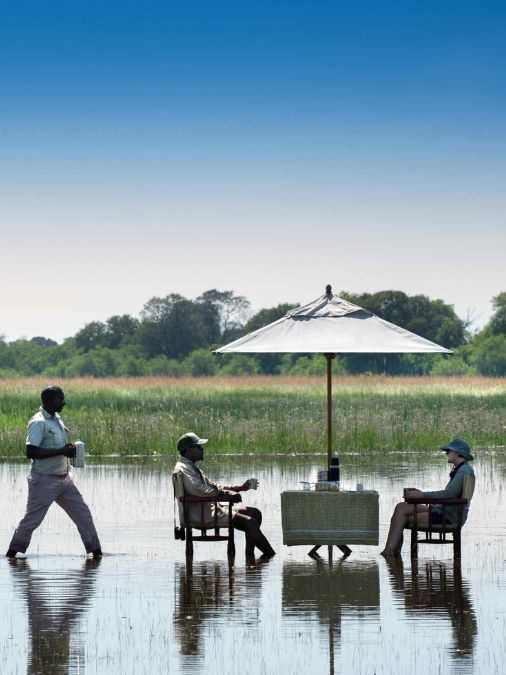Two people sitting at a table in a shallow river with a waiter approaching