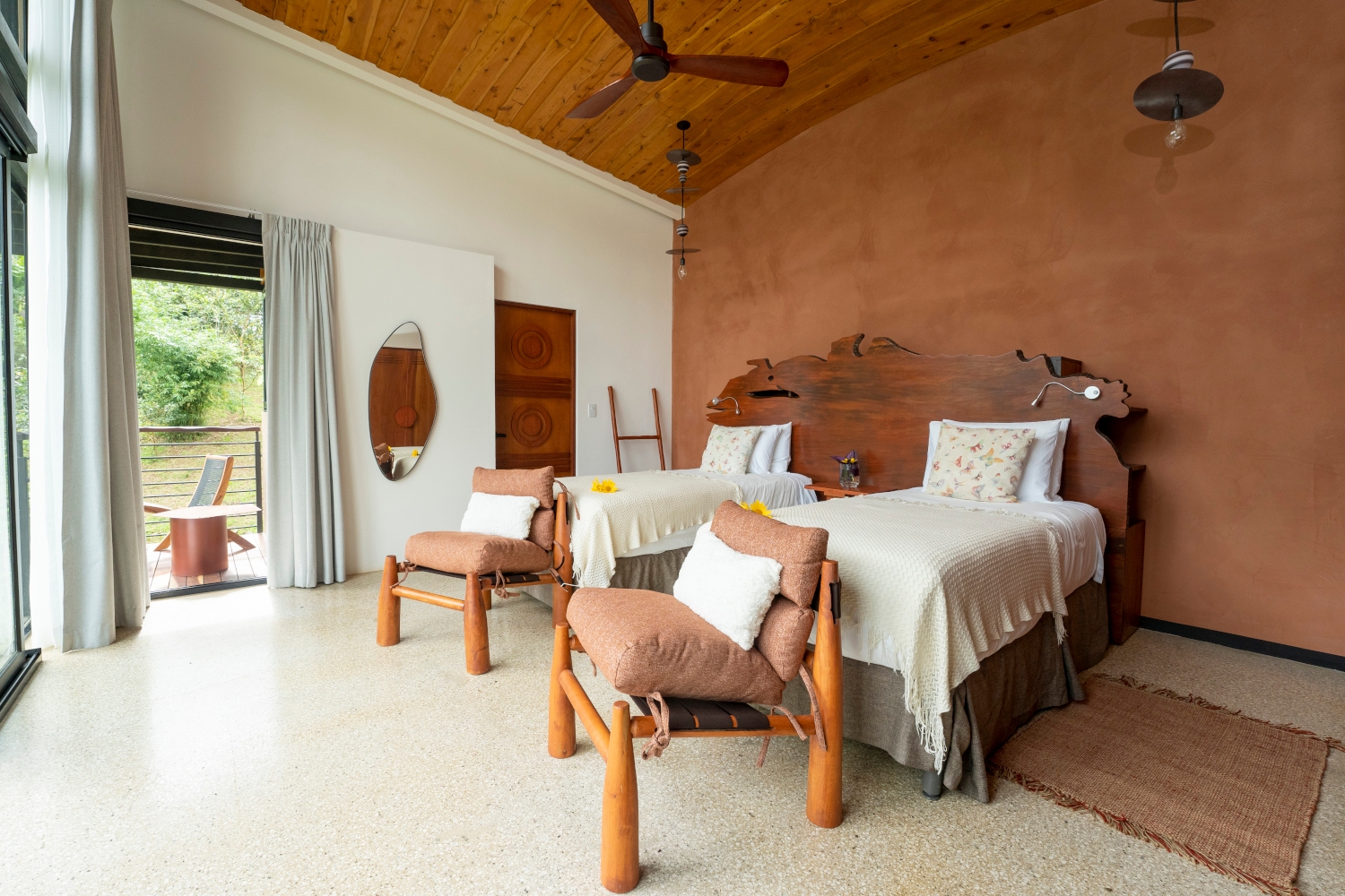 Interior of a bedroom with two beds with white sheets, brown wood walls, two wooden chairs chairs in front of the beds and a ceiling fan overhead.