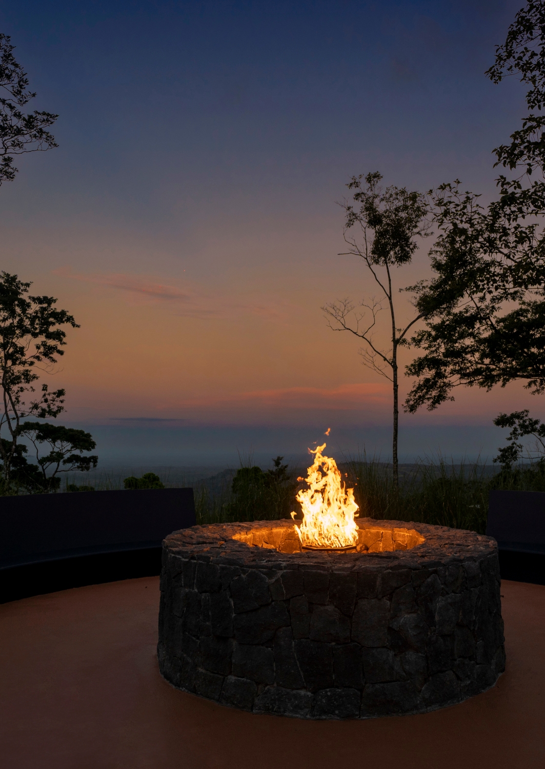 Lit firepit on a high terrace overlooking a darkening sunset sky with a few trees in the foreground.