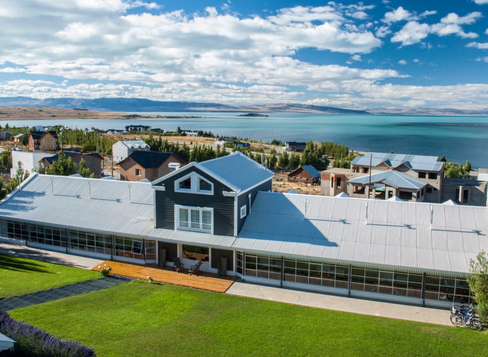 Aerial shot of the hotel with a green lawn in the foreground and scattered houses and trees in the background and the lake and shoreline beyond.