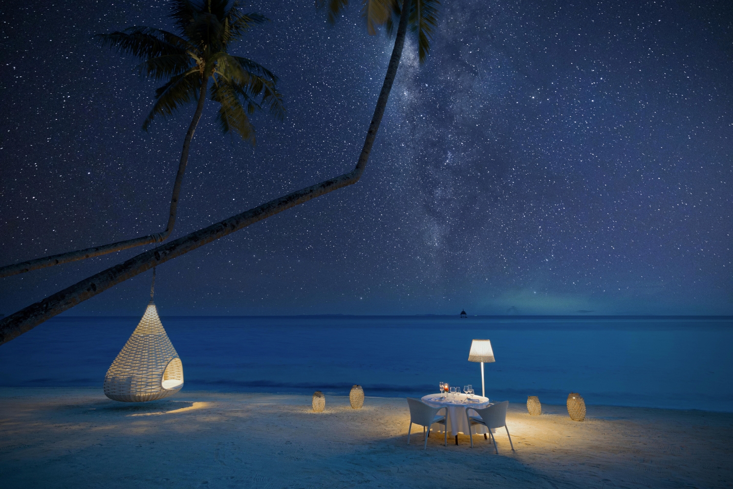 Starry sky with Milky Way over a beach table lit by a soft lamp next to the dark sea.