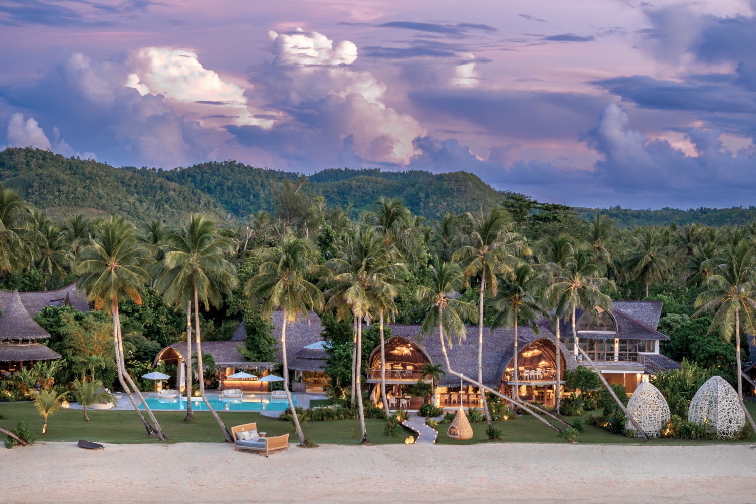 Distant shot of the Nay Palad Hideaway resort buildings set on the beach interspersed with tall palm trees and green mountains in the background.
