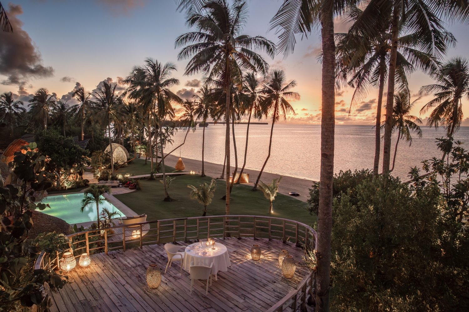 Wooden outdoor deck with tables surrounded by tall palm trees, a lawn and the beach and sea beyond.