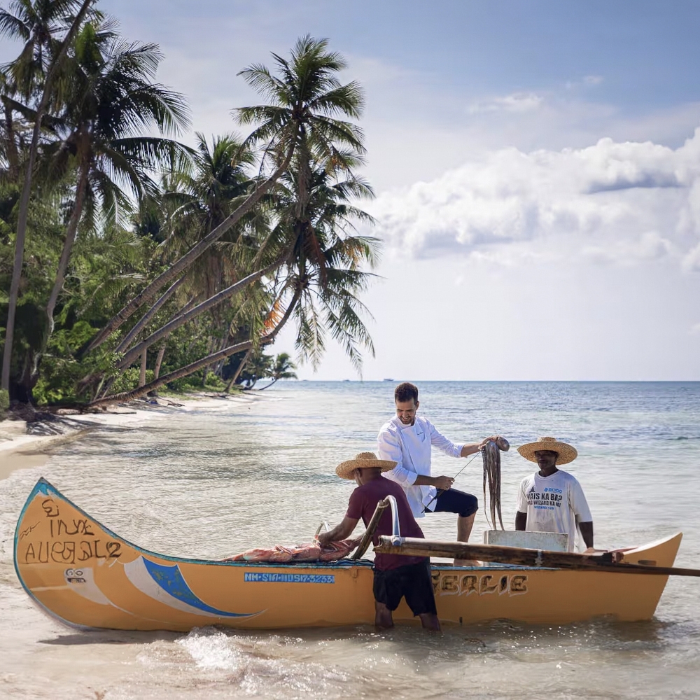 Guest and guides in a yellow canoe set on a beach of white sand, surrounded by shallow clear water and palm trees in the background.