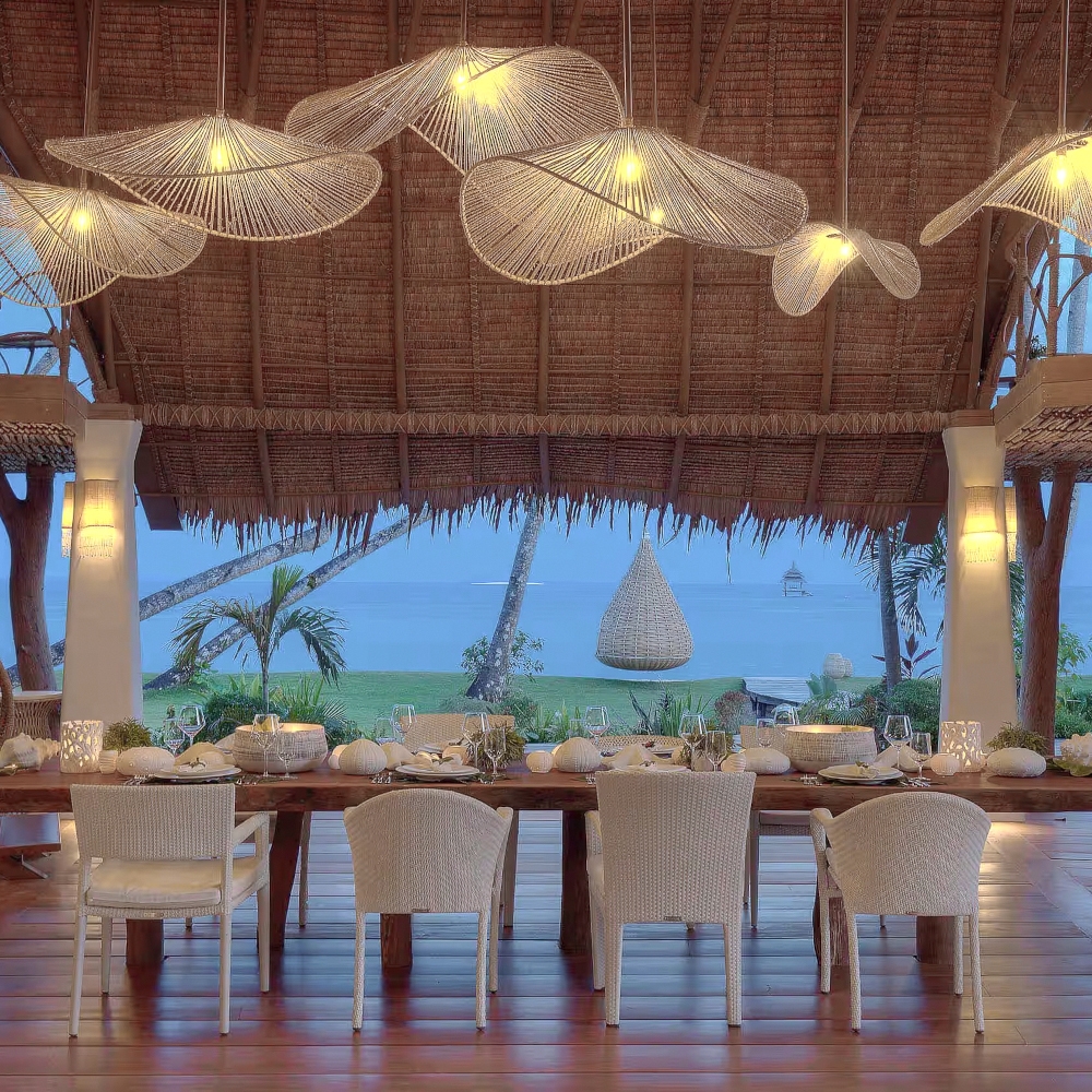 Dining table with white chairs set in an open-air wood deck with thatched roof and long weblike light fixtures overhead, with the sea and dark blue sky beyond.