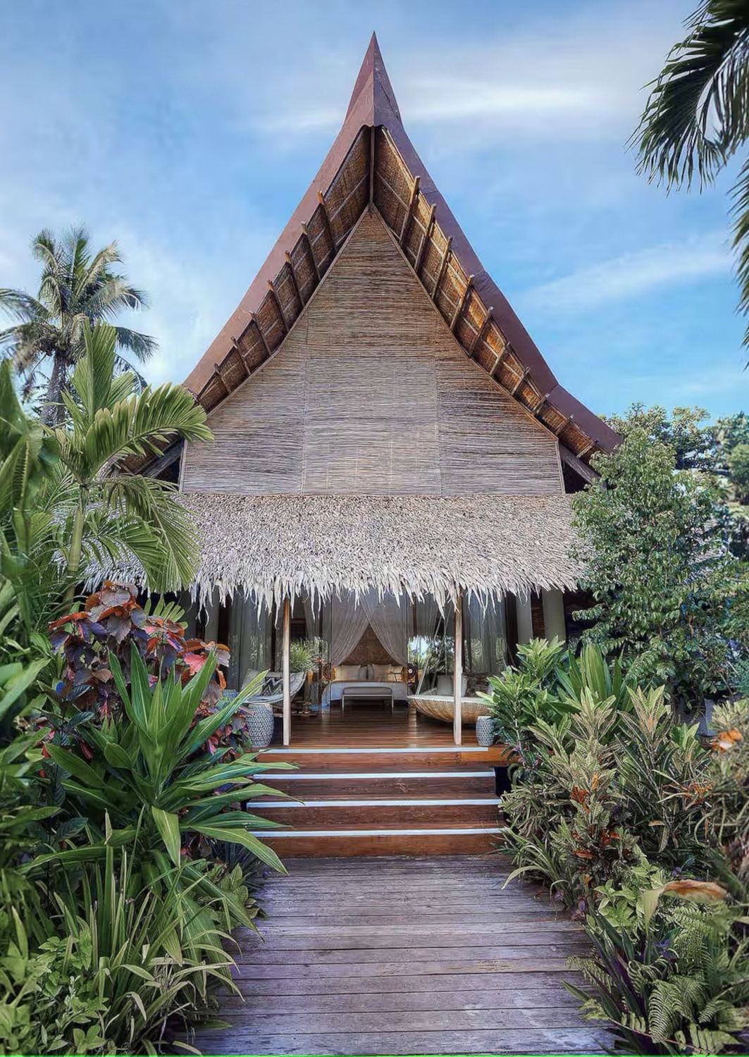 Entrance to a very sharply pitched roof thatched cottage, with steps leading up to an open-air bedroom, surrounded by green plants and palms.