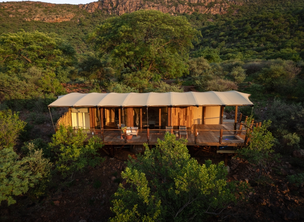 Exterior shot of a cliffside tented suite with canvas roof and wraparound wooden deck set among green trees with mountains in the distance.