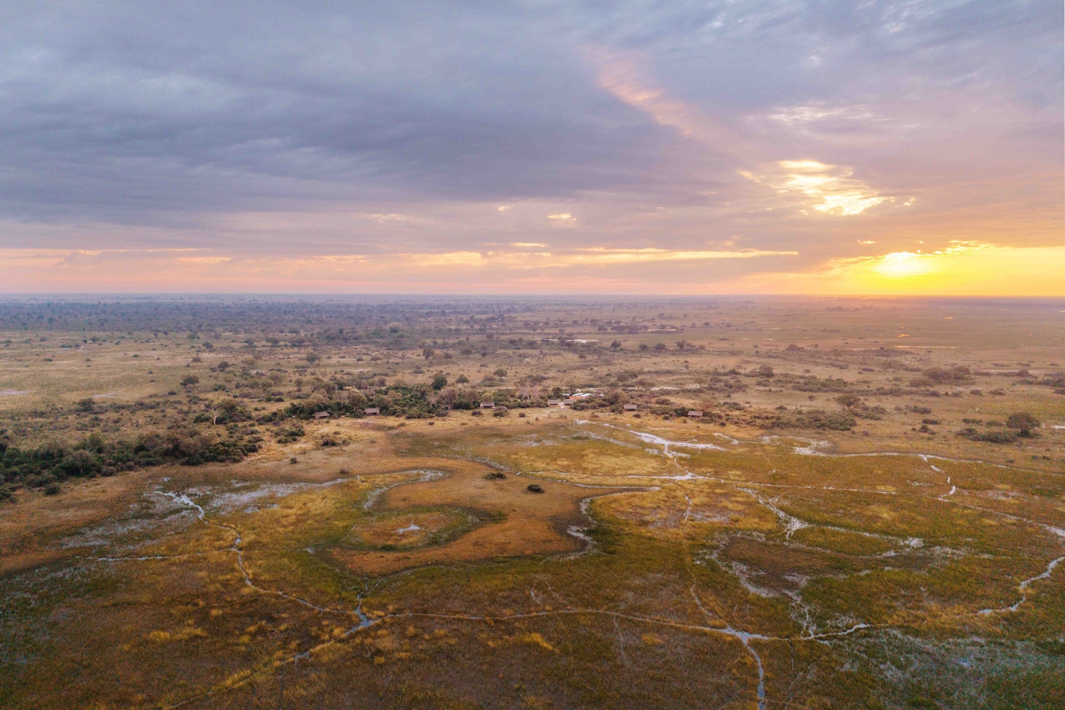 Aerial view of a vast green and brown plain of flooding and wetlands with sunset and clouds above.