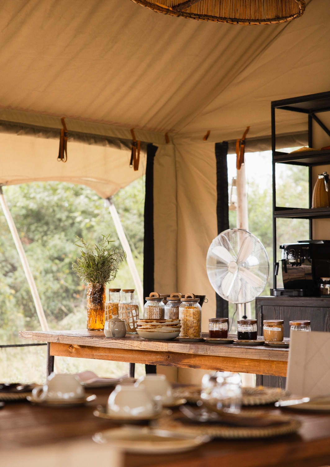 A table with a breakfast buffet of juices, cereals and foods inside a tented canvas lodge with open windows behind.
