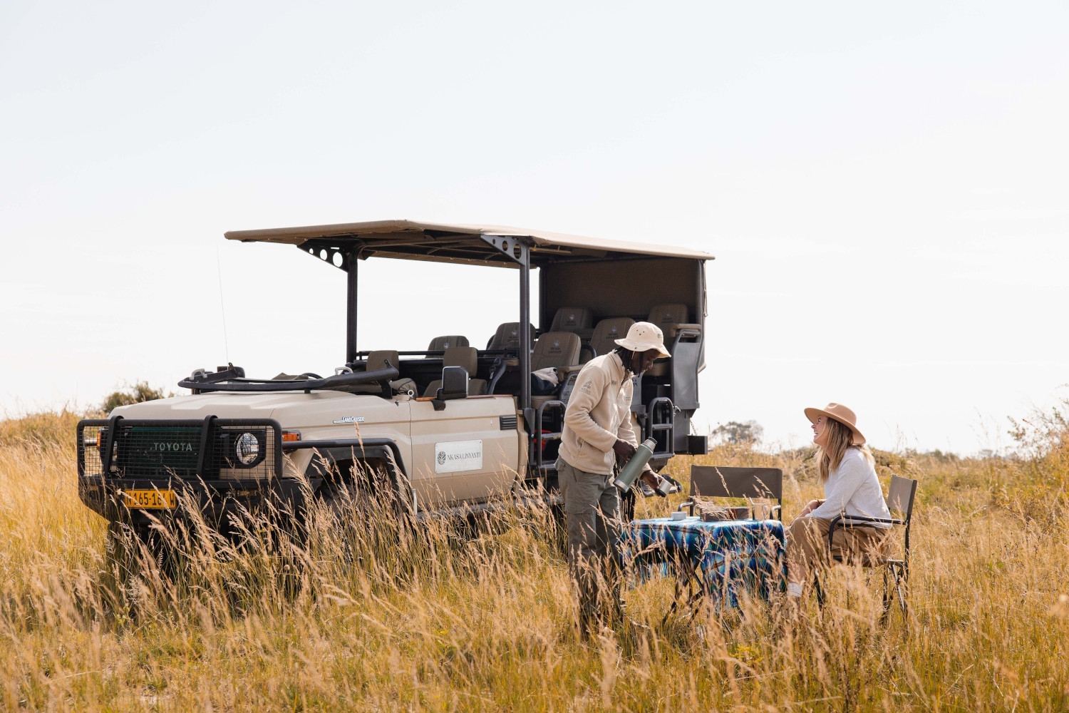 A safari guide stands next to a woman seated at a table in the bush in tall grass, with an open-sided safari vehicle in the background.