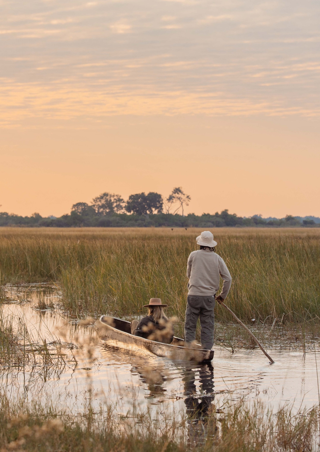 A person seated in a canoe being rowed by a guide standing up with a stick, gliding through water and reeds with an orange sunset sky.