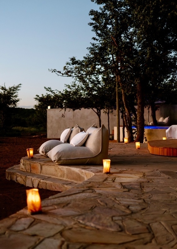 Two large floor pillows with smaller cushions on a flagstone patio, surrounded by lit luminarias, under a twilight sky.