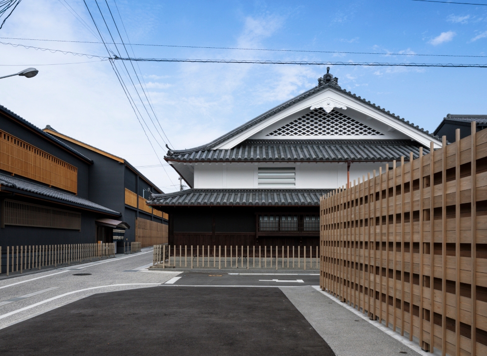 Exterior of a traditional Japanese building with white walls, pitched roof and tiled roof and a light wood fence around the right side.
