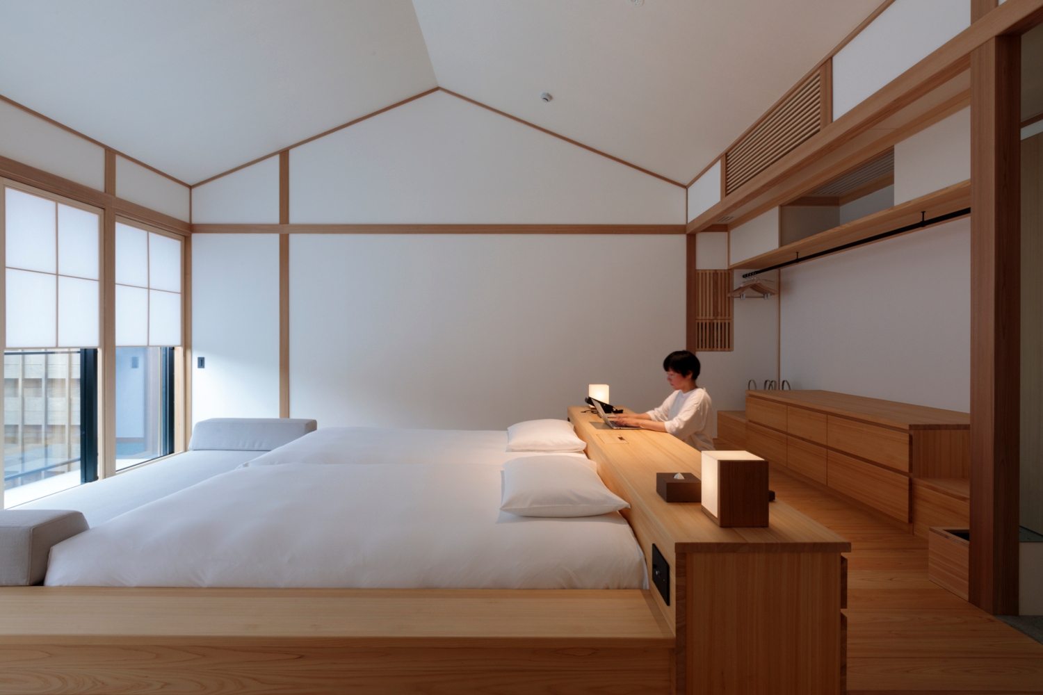 Interior of a bedroom with a minimalist bed with white bedding, light wood frame and white walls with light wood edging and accents, and a member of staff adjusting a lamp.